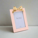 A soft pink photo frame with a gold ribbon detail at the top, designed to hold a 4" x 6" picture.